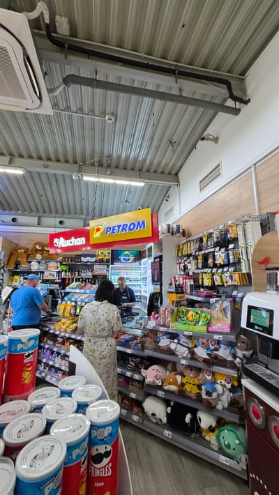 This media file captures an interior view of a retail store, likely a gas station convenience store, in Lehliu Gara, Romania. The perspective is slightly tilted, adding a dynamic feel to the scene.

The main subjects are several people, appearing to be shoppers and possibly staff, interacting within the store. One person in a blue shirt and cap is at a counter, while another, a woman in a floral dress, is browsing shelves. The shelves are stocked with a variety of merchandise, including Pringles snack cans prominently displayed in the foreground, stuffed animals, and other packaged goods. A large yellow and red "PETROM" sign is visible, indicating the store's affiliation with the Petrom gas station brand. Another sign reads "Auchan," suggesting a potential partnership or a store within a larger Auchan complex.

The setting is a brightly lit retail environment with corrugated metal walls and ceiling, typical of some service station stores. Fluorescent lighting illuminates the aisles. The presence of security cameras suggests a focus on safety and monitoring. The display of Pringles with text in Romanian ("SCANEAZA", "JOACA", "SARE") confirms the location.

The activity appears to be typical retail shopping. There are no clear indicators of the time of day or weather conditions. The overall atmosphere seems neutral, focused on commercial activity. The visible text includes brand names like "PETROM," "Auchan," "Pringles," and "BIC," along with promotional phrases in Romanian.