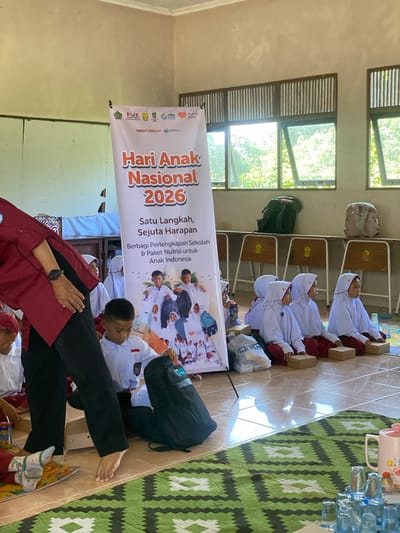 In a classroom setting in Handil Baru, Indonesia, a group of young students, predominantly girls in white hijabs and maroon skirts, are seated on the floor. They are facing a large banner that reads "Hari Anak Nasional 2026" (National Children's Day 2026) and other Indonesian text related to school supplies and nutrition. A woman in a maroon shirt and black pants, identified as part of the event, is standing and interacting with the children. Some children are looking towards her, while others are engaged with items in front of them, such as boxes or backpacks. The room appears to be a school, with wooden benches along the wall and open windows revealing greenery outside. The floor is tiled and decorated with a green and white patterned rug. Several backpacks and bags are placed on the benches and the floor. The scene suggests an event or distribution related to National Children's Day.