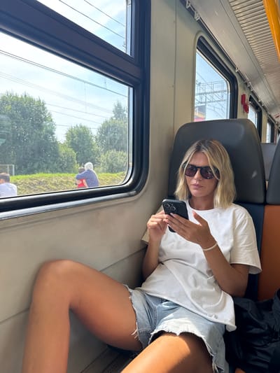 The image shows a woman in a train car, looking at her smartphone. She has blonde hair and is wearing sunglasses, a white t-shirt, and denim shorts. The train appears to be moving, as indicated by the blurred scenery outside the window, which includes trees and power lines. In the distance, two figures can be seen outside the train, one hugging the other. The interior of the train car shows a row of seats, some of which are visible. The woman is seated and appears relaxed. The location is identified as Tatarki, Russia.