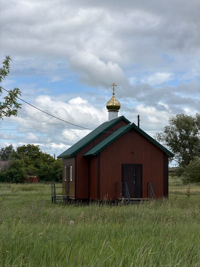 The image depicts a small, brown, wooden chapel with a green roof, situated in a grassy field. A golden dome with a cross tops the structure. The chapel has two doors and two windows. In the background, trees and a partially visible red brick structure can be seen. The sky is cloudy. The scene is located in the village of Bogdashkino, Russia.