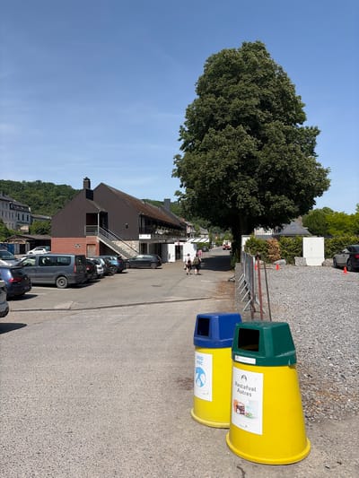 The image shows a street scene in Dinant, Belgium. Two large, yellow recycling bins with blue and green tops stand on a paved surface, likely a parking area. The bins have labels in French; one reads "Restafval Autre" and the other reads "PMD PMC." Behind the bins, a low metal barrier runs along the edge of the parking area. Cars are parked along the side of the street, adjacent to buildings with brown roofs and lighter-colored facades. A large tree stands to the left of the building, partially obscuring the view. In the background, more buildings and greenery can be seen. Two people are walking along the street further in the background. The sky is clear and blue.