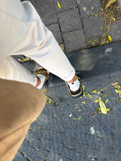 The image depicts a high-angle, downward view of the lower legs and feet of two individuals situated in West Jerusalem, Israel. The primary subject, centrally positioned, wears light beige trousers with rolled cuffs. This individual has white socks on both feet; the right sock displays a small black heart emblem on the outer ankle area. Footwear consists of a tan two-strap sandal with a gold-colored buckle on the left foot, and a dark brown two-strap sandal on the right foot. A second individual is partially visible in the lower left, wearing light brown trousers. The ground surface is composed of dark grey, square cobblestones, with a narrow strip of darker asphalt or a curb running along the right edge. Scattered across the cobblestones and asphalt are numerous fallen leaves, primarily yellow and light green, along with dry grass and miscellaneous debris. A small amount of white residue is visible on the cobblestones in the lower right portion of the frame.