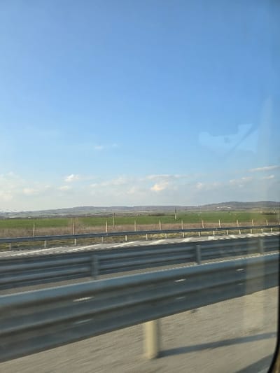 The image depicts an exterior view from a moving vehicle, likely a car, situated on a multi-lane highway near Rupkite, Bulgaria, under a clear blue sky with sparse white cumulus clouds. In the foreground, a multi-beam galvanized metal guardrail runs horizontally across the frame, partially obscuring the asphalt road surface which exhibits motion blur. Visible behind the guardrail are white dashed lane markings on the road. Beyond the road, cultivated green fields extend into the mid-ground, delineated by a series of vertical wooden or metal posts. The distant horizon reveals undulating terrain with faint tree lines or vegetation. A faint, irregular reflection, possibly of an interior vehicle component, is present on the window surface in the upper right quadrant of the image.