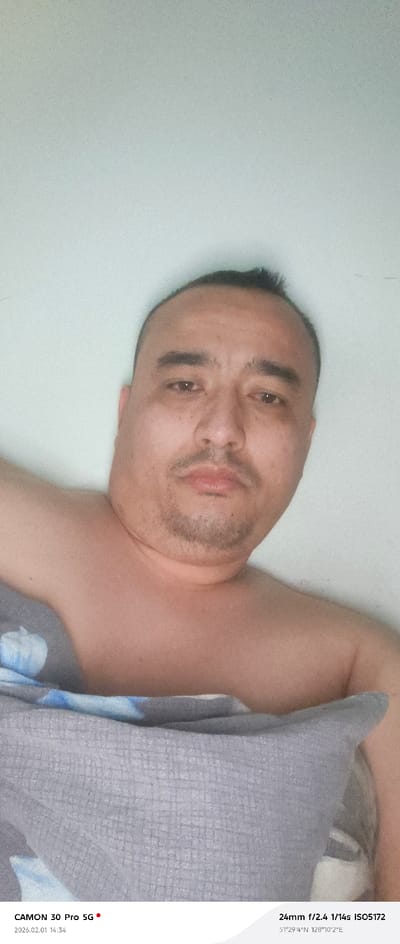 An up-close selfie featuring a man with short, dark hair and a trimmed mustache and goatee. He appears to be partially covered by a gray patterned blanket or pillow. The background is a plain, light-colored wall. The camera settings are visible at the bottom, noting a CAMON 30 Pro 5G, focal length of 24mm, aperture f/2.4, shutter speed of 1/14s, and ISO 5172. The date and time are 2026.02.01 14:34, with the location indicated as 51°29'4"N 128°10'2"E.