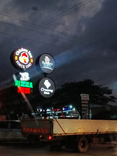 The image captures a commercial street scene at dusk or night in Marondera, Zimbabwe, characterized by illuminated business signs and a dark, cloudy sky.

In the foreground, four circular, brightly lit signs are stacked vertically, advertising food establishments. From top to bottom (assuming correct image orientation), the signs display:
1. "BULLES INN" with a distinctive crown and lion logo.
2. "Creamy Inn" featuring an ice cream cone graphic.
3. "Chicken Inn" accompanied by a chicken head logo and the text "SINCE 1987".
4. A vibrant sign with the word "PIZZA" in stylized green and red lettering.

Power lines are faintly visible against the dark, overcast sky on the left side of the frame. On the right side of the image, a large white box truck, possibly a delivery vehicle, is either parked or moving, with its rear section and a visible tire. In the background, further to the right, a tall roadside sign pole lists multiple businesses, including "We love", "PHARMACY", "GROCERIES", "SERVICES", and "STORES", though some text is blurry due to distance and lighting. Distant lights from other establishments are scattered in the background, reinforcing the commercial setting.

The scene depicts a typical evening in a commercial district, with businesses open and their signs illuminated. No people are visible. The time of day is clearly evening or night, indicated by the dark sky and reliance on artificial lighting. The atmosphere is functional, typical of a bustling commercial area after dark.