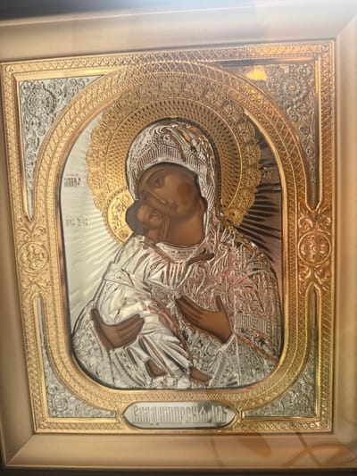 The media file displays a framed religious icon, likely of the Virgin Mary and Christ Child, rendered in embossed metal with gold and silver finishes. The central figures are depicted in profile, with the Virgin Mary embracing the child. Their faces and hands are rendered in a darker, flesh-toned metal, contrasting with the ornate, highly detailed silver and gold metallic robes and halos. The background behind the figures features radiating lines, suggesting a divine light. The icon is encased in a decorative frame, with intricate gold detailing on the inner border. The outer frame is a simpler, golden rectangular border. The overall aesthetic suggests a traditional Orthodox Christian icon, possibly a representation of the Hodegetria icon type. There is some Cyrillic text visible vertically on the right side of the icon, but it is partially obscured and difficult to read clearly. The lighting in the image suggests an indoor setting, possibly a church or a home, with a soft light source casting a slight glare on the metallic surfaces. The image is oriented horizontally. The location context of Николо-Урюпино, Russia, reinforces the likely religious and cultural significance of the object.