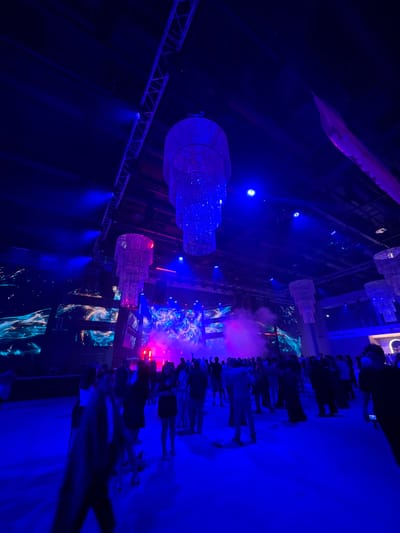 The image depicts a large indoor event space illuminated with vibrant blue and purple lights. Multiple tiered chandeliers, covered in reflective material, hang from the dark ceiling, refracting the light and adding to the festive atmosphere. Large screens in the background display dynamic, abstract visuals with swirling colors and textures. A haze of pink and purple smoke drifts through the space, partially obscuring the view of the crowd. Silhouettes of numerous people are visible, scattered across the floor, suggesting a gathering or party. The overall impression is one of a lively, modern event. There is no specific visual information in the image to suggest the location is in Akhalubani, Georgia.