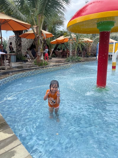 The image depicts an outdoor water park scene in Juwana, Indonesia. A young child, female, approximately 5 years old, stands mid-torso deep in a light blue tiled swimming pool. She wears an orange short-sleeved shirt and light-colored shorts, with a blue wristband on her right wrist. Her dark hair is wet. She faces forward, smiling, with both hands raised, each displaying a V-sign with the index and middle fingers.
To the child's right, a prominent red pole with a red and yellow mushroom-shaped top functions as a water spray feature, emitting streams of water into the pool, causing splashes. Additional yellow and blue water play structures are partially visible in the distant right background.
The pool's edge is a light blue tile, bordered by a checkerboard pattern of dark blue and white tiles. Beyond the pool, a paved area leads to multiple orange patio umbrellas providing shade over wooden tables and chairs. Several individuals are seated at these tables, and others are visible standing or walking in the background. Multiple tall palm trees are interspersed throughout this area. Two thatched-roof huts are situated behind the seating arrangements. The sky above is overcast. No direct interaction between the child and other individuals is depicted; the child is the focal subject.