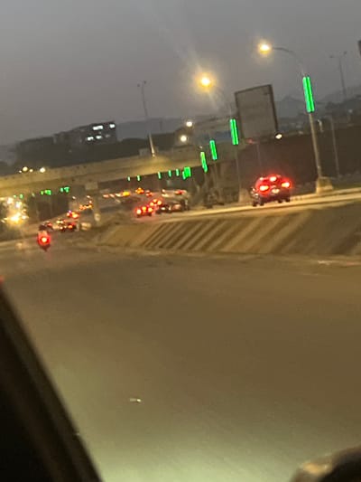 The image depicts a nighttime scene, possibly taken from a high vantage point, showing a road with traffic in Wupa, Nigeria. The road is illuminated by streetlights, with cars displaying red taillights indicating they are moving away from the viewer or stopped. Green lights are visible above the road, which may be from the streetlights on the side of the highway. The environment is hazy or foggy, reducing visibility. A portion of the interior frame where the image was taken is visible in the bottom right.
