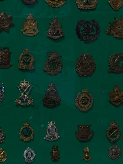 A collection of metal military badges is displayed on a green background. The badges vary in design and color, ranging from bronze and silver to darker tones. Some depict crowns, wreaths, eagles, and crossed swords, suggesting different military units or ranks. They are arranged in a grid pattern, and there are many more badges shown in the image.