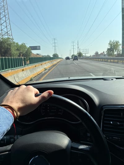 A person is driving a car on a highway, looking out the front windshield, driving on a highway next to a concrete divider wall. There is green foliage on the other side of the wall. Power lines and electrical towers are visible in the distance. The location is Ciudad López Mateos, Mexico. The car's dashboard and steering wheel are visible. The driver's hand is visible, holding the steering wheel, and the driver is wearing a light blue shirt. The car's dashboard displays a time of 10:24:18 and a mileage of 166633 km.