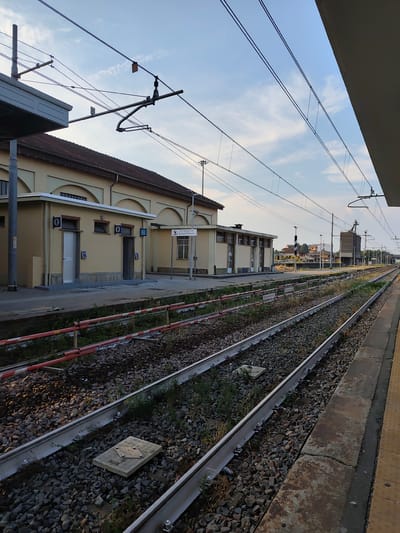 The image depicts a train station platform in Carmagnola, Italy. The scene is viewed from the platform, looking down multiple train tracks. On the left, a light-yellow building with arched windows and a covered walkway is visible. Adjacent to this building is a smaller, also light-yellow structure with two doors, each marked with a male and female restroom icon. Beyond the tracks, on the right, the platform continues with a yellow-striped edge. Overhead, numerous wires and support structures for the overhead catenary system are present, indicating an electrified railway line. The sky above is a light blue with scattered white clouds. The railway tracks are laid on a bed of ballast, with some vegetation growing between the ties. In the distance, more buildings and what appear to be silos are visible. The lighting suggests it is either early morning or late afternoon, with the sun casting long shadows. There are no people visible in the frame.