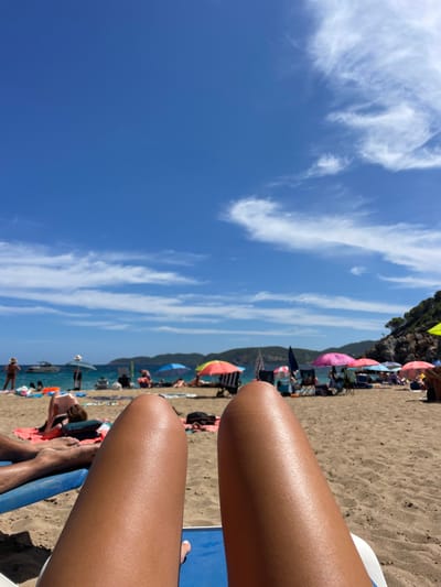 The image presents a first-person perspective from a beach lounger on the sandy shores of Cala De Sant Vicent, Spain, under a clear blue sky with scattered white cirrus clouds. The foreground is dominated by the tanned legs of an individual resting on a blue and white beach lounger.
The midground reveals a populated beach scene. Numerous individuals are visible, most engaged in relaxation, sunbathing, or sitting under a variety of colorful beach umbrellas, including shades of pink, orange, green, and striped patterns. Beach towels, bags, and portable chairs are scattered across the beige sand. To the left, a person is partially visible lying on an orange towel. Near the water's edge, several people are standing or wading in the turquoise-blue sea. A white motorboat is anchored in the water towards the left.
The background features the calm, blue waters of the cove, extending towards distant, verdant hills and rocky outcrops that define the bay. The overall environment indicates a sunny day at a busy coastal resort location.