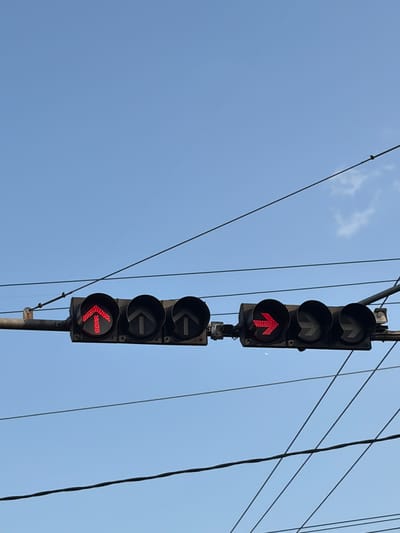 The image shows a traffic light with red arrows pointing straight up and to the right. The traffic light is suspended over an empty street, with power lines crisscrossing the sky above. The sky is a clear blue, with a few wispy clouds. The traffic light is positioned at an intersection, and the red arrows indicate that traffic is currently stopped. The image is taken from a low angle, looking up at the traffic light. There are no people or vehicles visible in the image. The scene appears to be quiet and still. The presence of power lines suggests an urban or suburban environment. The specific location in the city of Magelang, Indonesia is not identifiable from the image alone, as traffic lights and power lines are common in many urban areas.