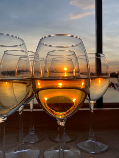 The image shows a row of wine glasses partially filled with a light-colored liquid, likely white wine, set against a backdrop of a twilight or sunset sky. The glasses are arranged horizontally, and the light from the setting sun is reflected in the wine, creating a golden hue. The glasses are clean and transparent. In the background, a dark horizontal line may represent a railing or a structural element. The setting suggests an outdoor location, possibly a restaurant or a bar with a view. The exact location within the city of Aramil, Russia, cannot be determined from the visual data.