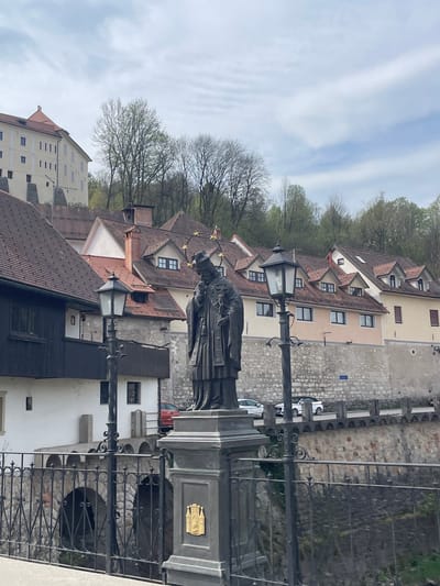 The image depicts a bronze statue of Saint John of Nepomuk situated on a bridge in Skofja Loka, Slovenia. The statue is positioned in the middle ground, with a backdrop of buildings featuring red-tiled roofs and light-colored facades. A wrought-iron fence is visible in the foreground. The sky is overcast with wispy clouds. The buildings indicate a historic urban environment.