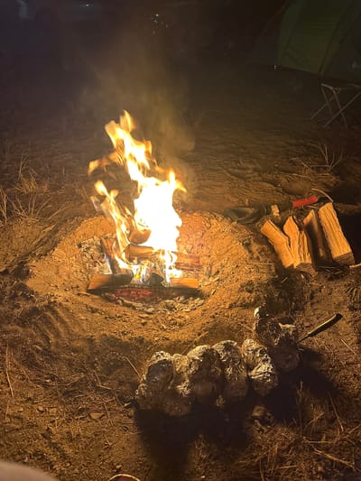 A campfire burns brightly in a dirt pit at night. The flames are orange and yellow, casting a warm glow on the surrounding dry grass and soil. To the left of the fire, a green tent is partially visible, suggesting a camping or outdoor setting. To the right of the fire, a pile of objects wrapped in aluminum foil are arranged on the ground, likely food being cooked in the embers. A shovel with a red handle and a metal blade rests near a stack of chopped firewood, indicating preparation for the fire. A knife is also stuck into the ground near the foil-wrapped items. The scene is dimly lit, characteristic of nighttime. The overall mood is cozy and rustic, evoking a sense of outdoor adventure. There is no visible text in the image.