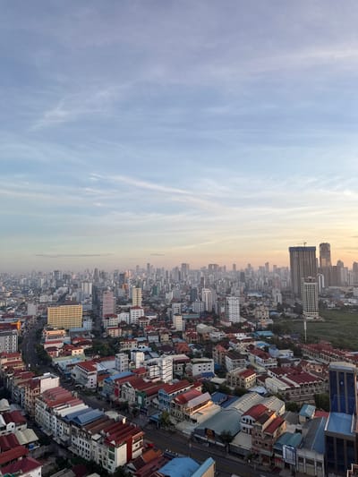 This is a high-angle, panoramic view of a dense urban landscape in Khan Mean Chey, Cambodia. The image is oriented vertically, with the sky dominating the left half and the city filling the right. The sky is a pale blue with wispy white clouds, suggesting it's either early morning or late afternoon. The sun is low on the horizon, casting a warm, golden glow on the cityscape, particularly on the buildings closest to the horizon.

The city is a sprawling mix of residential and commercial buildings, with a variety of architectural styles and roof colors, including prominent red and blue tile roofs. Many buildings appear to be mid-rise, with some taller modern structures in the distance, including one under construction with a visible crane. The density of buildings is striking, with narrow streets visible between them. There are also areas of greenery interspersed within the urban fabric.

No people are clearly visible, suggesting the photo was taken from a significant height. The overall impression is one of a busy, developing city bathed in the soft light of dawn or dusk. The visual cues of dense housing, varied building heights, and the presence of construction indicate an active and growing urban environment. There is some faint text visible on signs on some buildings, but it is largely illegible due to the distance and resolution.