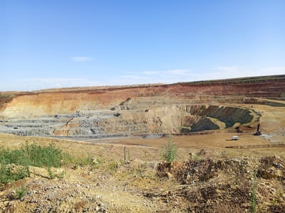 The image shows a large open-pit mine under a clear blue sky. The mine has numerous terraced levels exposing layers of earth in various shades of brown, orange, and gray. Several pieces of heavy machinery, including excavators and trucks, are visible on the different levels of the mine, indicating ongoing operations. In the foreground, the terrain is rough and covered with rocks and sparse vegetation. The scene suggests an industrial or mining landscape. There are no people clearly visible in the image. The location is identified as a mine in Raygorodok, Kazakhstan.