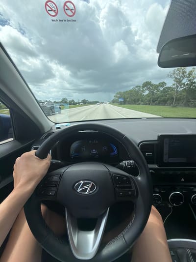A first-person perspective shot from inside a car shows the driver's hands on the steering wheel, looking forward onto an overcast highway. The driver's left hand is visible gripping the steering wheel, with their legs also in view. The dashboard displays the speed as 74 mph and the car's lane assist system is active. A digital screen on the right shows media controls. Two "no smoking" signs are affixed to the windshield, with the text "Help Us Keep Our Cars Clean" below them. Outside, trees line the highway, and other vehicles are visible on the road. The scene appears to be in Vero Beach, Florida, United States, given the presence of a "Parking Permit" sticker on the windshield, a common sight in many Florida cities.