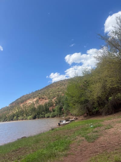 The image depicts a daytime outdoor scene at Ait Ouaarda, Morocco, featuring a body of water adjacent to a vegetated hillside. A clear blue sky with scattered white cumulus clouds dominates the upper portion. A large, still body of brownish-grey water occupies the left side, extending to the lower-left corner. The immediate foreground consists of a dirt bank with patches of green grass and scattered litter, including a white plastic bag. A small, light-colored open boat with two outboard motors is partially beached on the muddy shore near the right-center, with a white object visible on or near it. To the right, a dense cluster of trees with light green foliage lines the bank, casting some shade. On the left, a steep hillside rises from the water, displaying exposed reddish-brown earth interspersed with green shrubs and coniferous trees, some appearing sparse or dry. No individuals are visible.
