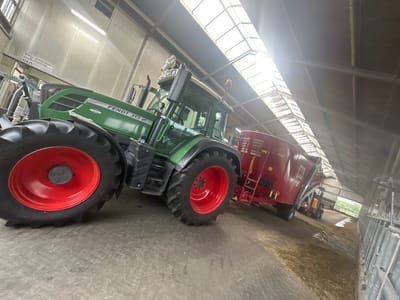 The image depicts a large green Fendt 313 Vario tractor parked inside a barn or agricultural building. The tractor is positioned at an angle, with its large rear tire, featuring a red rim, prominently in the foreground. The building has a high ceiling with natural light filtering in through a series of skylights. To the right of the tractor is a large red trailer or feed mixer. In the background, a smaller orange forklift is visible, and beyond that, an opening shows a glimpse of the outdoors, suggesting a rural setting. A sign with text, including "FENDT 313 Vario," is visible on the side of the tractor. Another sign with text that appears to be a company name and location ("Bosma Melktechniek St. Nicolaasga") is affixed to the wall in the upper left quadrant of the image. The floor of the building is concrete, with some scattered straw or animal feed. The overall impression is one of agricultural machinery stored within a functional farm building. The city of Wilhelminaoord, Netherlands, is a rural area known for its agricultural activities, making this scene consistent with the location.