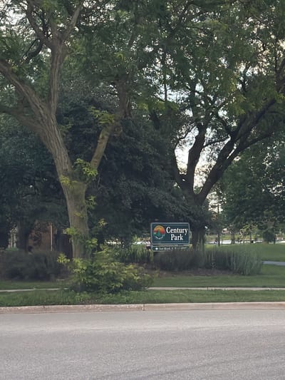 The image depicts a park entrance sign on a street in Vernon Hills, USA. The sign, oriented vertically, reads "Century Park" in large white letters against a dark green background. Below "Century Park," there's a circular logo with a sunrise or sunset motif, and further down, smaller text indicates "Ice Rink, S. Bike." The sign is set against a backdrop of lush green trees and foliage. To the right of the sign is a paved road and a sidewalk. The scene appears to be an exterior shot on a bright, possibly late afternoon or early morning, given the lighting and shadows. The overall impression is of a serene park entrance.