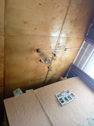An interior shot captures a room with a sloped, unfinished plywood ceiling, indicative of a rustic or unfinished dwelling. Dominating the center of the frame is a multi-tiered chandelier, its metallic structure adorned with decorative elements. A portion of a window with purple curtains is visible on the right, letting in diffused natural light. In the foreground, a light brown, textured surface, likely a mattress or covered bed, stretches across the bottom of the image. Placed on this surface is a white multi-photo frame containing several small pictures. Boxes are stacked in the upper left corner. The scene appears to be a room in a private residence in Churapcha, Russia, possibly a bedroom or living space.