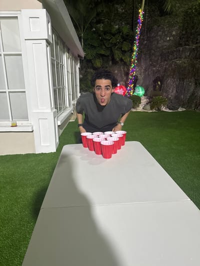 A playful, young man poses outdoors next to a beer pong setup. He's wearing a dark gray shirt, sports a watch, and sticks his tongue out at the camera. Behind him, a brightly lit window and festive lights are visible, hinting at a celebration. The beer pong table sits on artificial turf, with a rock formation and greenery in the background. The overall scene suggests a fun, casual outdoor party.