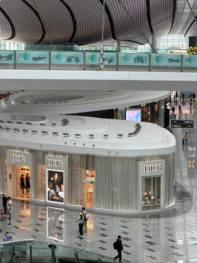 The image shows an interior view of a modern airport terminal, likely in Beijing Daxing International Airport in Guangyang District, China, given the architectural style and presence of Dior stores. The scene is characterized by its vast, open space with a high, curved ceiling structure made of ribbed panels. Natural light filters in from large windows and the reflective floor accentuates the bright, clean environment.

Prominently featured is a Dior boutique with its signature signage and a large display window showcasing handbags. Other commercial establishments are visible in the background, along with directional signs and advertising screens. People are scattered throughout the terminal, walking, standing, and interacting. Some are dressed casually, while others appear to be airport staff in uniforms. The floor is highly polished and reflects the light and overhead structures, creating a sense of depth and spaciousness. Artwork or informational displays are mounted on a wall in the upper part of the image. The overall impression is one of a busy, upscale, and contemporary transportation hub.