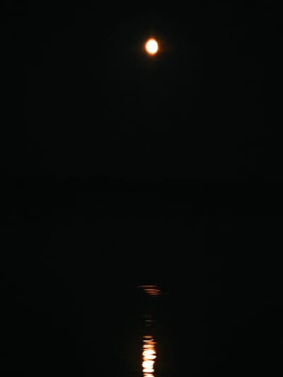 The image captures a night scene with a dark sky. A bright, circular object, possibly the moon or a distant light source, is visible in the upper portion of the frame. Below this, a reflection of the light source appears on a dark, still surface, likely water, creating shimmering streaks of light. The reflection is elongated and broken, suggesting ripples on the water. The scene is devoid of any human presence or man-made structures, conveying a sense of isolation and natural ambiance. No specific location in Shirokii Log, Russia, can be determined from the provided image.