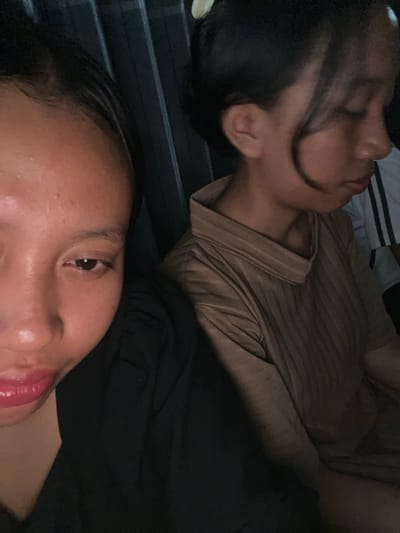 The image shows two individuals, a woman in the foreground and another woman in the background. The woman in the foreground is positioned to the left, her face close to the camera, with her right eye visible and looking towards the camera. She has dark hair and is wearing a black top. Her lips are visibly glossy and red. The woman in the background is on the right, her face turned to the side with her eyes closed. She has dark hair pulled back and is wearing a light brown, textured shirt. The background appears to be a dark, possibly interior space with vertical lines, suggesting a curtain or blinds. The image was taken in Jambearum, Indonesia.