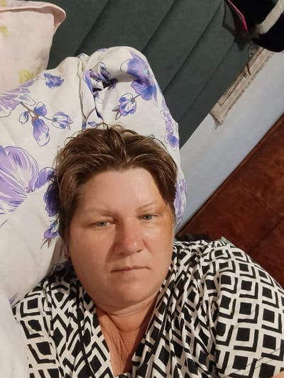A woman with short brown hair and blue eyes is lying on a bed, looking towards the camera. She is wearing a black and white patterned shirt. To her left is a pillow with a floral pattern. The background consists of a dark gray tufted headboard and a wooden surface on the right. The lighting appears to be indoor. There is no indication of the specific location within the city of Койлык, Kazakhstan.