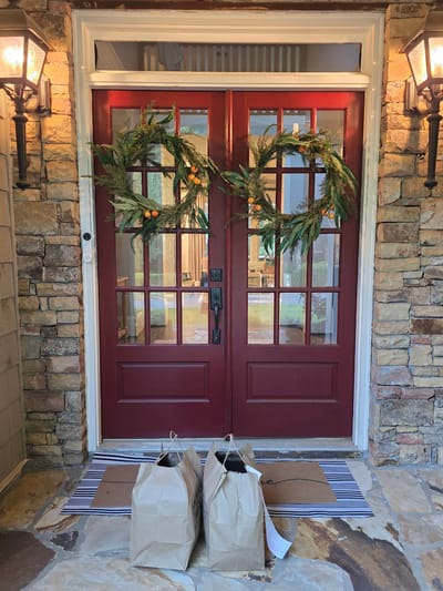A pair of red double doors, adorned with two circular wreaths made of greenery and small orange fruits, stands as the focal point of the image. The doors are set within a stone facade, flanked by two decorative lanterns mounted on the wall. In front of the doors, a striped doormat is partially covered by two brown paper grocery bags, suggesting a recent delivery. The scene is outdoors, on a stone porch.