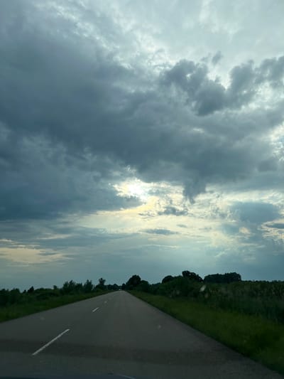 The image depicts a straight asphalt road extending into the distance through a rural or semi-rural landscape in Pohreby, Ukraine. The perspective is from within a vehicle, looking forward. The sky is largely overcast, dominated by dark grey and lighter grey cloud formations. A bright area on the upper right-mid portion of the sky indicates the sun partially breaking through the clouds, casting diffuse light and some visible light rays.
The two-lane road features white dashed lane markers. The immediate foreground shows the road surface and a slight dark reflection consistent with a vehicle's dashboard. On both sides of the road, dense green vegetation, including tall grass, bushes, and trees, lines the verges and fields. The vegetation on the right appears thicker and taller than on the left. In the far distance, a dark line of trees defines the horizon. A light-colored vehicle is faintly visible on the road in the far distance, receding from the viewer. No individuals are present in the visible portion of the scene. The lighting suggests an overcast condition, possibly during late afternoon or early morning, with the sun attempting to emerge from behind the clouds.