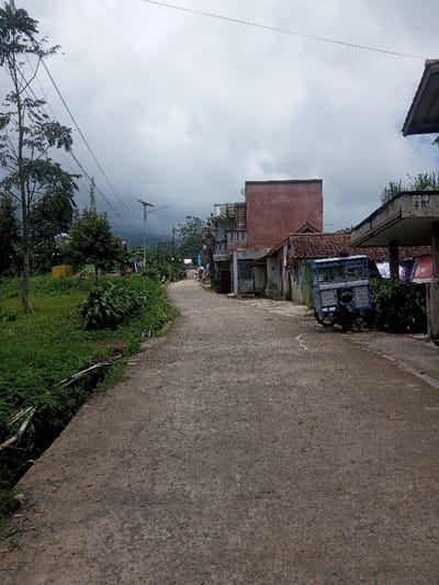 The image depicts an unpaved road in Ura-Dozhi, Bhutan, under a heavily overcast sky. The road, composed of dark gravel and dirt, extends from the foreground into the distance, appearing to curve slightly to the left. On the left side of the road, a green embankment is covered with dense vegetation, including a large tree with sparse foliage in the foreground and various bushes and smaller trees extending into the midground. A narrow ditch or drainage channel runs along the immediate left edge of the road. Utility poles with multiple overhead wires span the scene, and a solar-powered streetlamp is visible further down the road on the left.
On the right side of the road, a series of structures line the thoroughfare. In the immediate foreground on the right, a portion of a concrete overhang is visible, with laundry items hanging from what appears to be a clothesline. Adjacent to this, a dark-colored motorcycle is parked next to a blue and white enclosed cart-like structure. Further along, a building with a reddish-brown tiled roof is present, followed by a prominent, rectangular, reddish-brown two-story building with a flat top. Additional buildings, varying in construction and condition, are visible further down the road on the right. No individuals are present in the frame. Distant hills or mountains are partially obscured by the cloudy sky in the background.