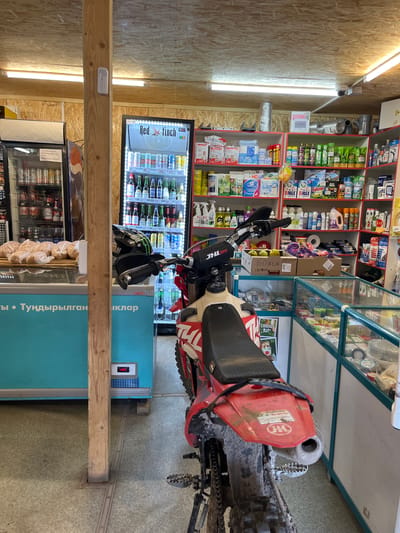 The image depicts the interior of a small store located in Krasnyi Yar, Russia. A red and white dirt bike is parked in the foreground, angled towards the right side of the frame. The store is stocked with various goods, including drinks in a refrigerated display case on the left, and shelves lined with cleaning supplies, hygiene products, and other household items on the right. A freezer chest is visible behind the dirt bike's handlebars, and a wooden pillar stands prominently in the center of the image. The floor is covered with light-colored linoleum. The overall impression is of a functional, well-stocked convenience store.