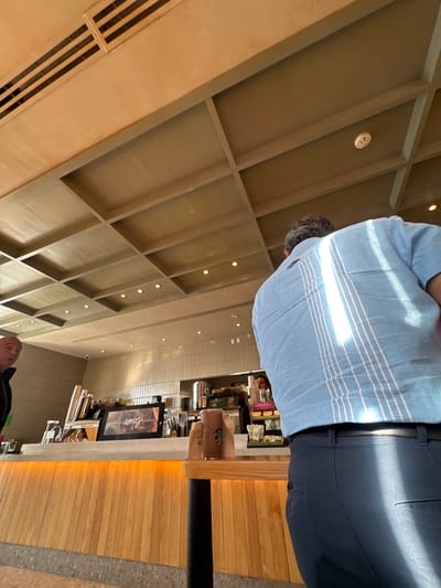 The image shows the interior of a Starbucks in Santa Ana Xalmimilulco, Mexico. A person wearing a light blue striped shirt and dark pants is seen from behind, possibly standing at the counter. The Starbucks counter is made of light-colored wood paneling. A digital menu board displays an image of a beverage. Behind the counter, there are coffee machines, beverage ingredients, and cups. Another person is visible towards the bottom of the image, and another person is visible behind the counter. The ceiling has a grid-like structure with recessed lighting.