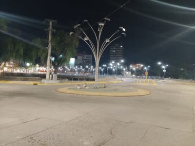 This is a nighttime shot of a roundabout in Villa Carlos Paz, Argentina. The roundabout is constructed of concrete and has a yellow painted border. Inside the roundabout is a light fixture shaped like a tree, made of silver metal, and surrounded by large, round stones. There are street lights and buildings visible in the background, as well as trees to the left of the roundabout. There are also traffic cones visible on one side of the roundabout. The foreground is mostly concrete road surface.