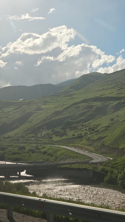 This image depicts a mountain road in Razv, Russia, with a steep, grassy slope on one side and a turbulent river on the other. The road, which curves sharply, appears to be a mountain pass, as evidenced by the dramatic elevation changes and the winding nature of the asphalt. The river below is wide and fast-flowing, with a rocky bed and glistening water, suggesting recent rainfall or snowmelt. The overall scene is one of rugged natural beauty, with the vibrant green of the mountains contrasting with the grey of the river and the blue of the sky.

The time of day appears to be late afternoon, given the warm, directional sunlight casting long shadows across the landscape. The weather seems fair, with scattered white clouds dotting the bright blue sky. The presence of a guardrail in the foreground indicates that this is a publicly accessible road, likely a tourist route or a route used for transportation through the mountainous terrain. There are no people visible in the image, but the presence of the road and guardrail suggests human activity in the area. The vastness of the landscape and the dynamic flow of the river create a sense of awe and the power of nature.