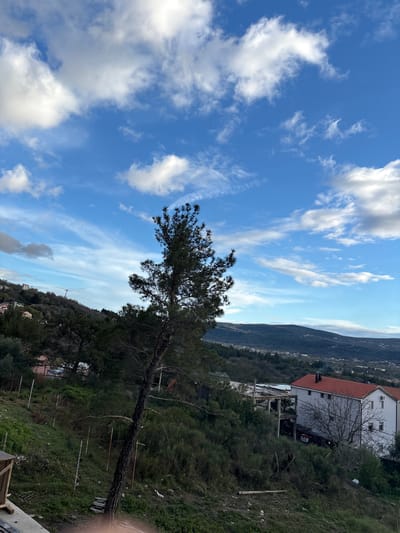 The image depicts a panoramic view of the Kavač region in Montenegro, characterized by a mix of natural landscape and residential development under a partly cloudy blue sky.

In the foreground, a prominent, slender conifer tree with a dark trunk and green needles leans slightly to the viewer's left. The immediate ground slopes downwards, covered in green and dry vegetation, including shrubs and grasses. A rudimentary fence line is visible on this slope, consisting of several vertical, weathered metal posts with remnants of wire mesh. A wooden pallet and other minor debris are present on the ground. A partial wooden structure, possibly a construction element or shed, is visible in the bottom-left corner.

The midground features a hilly terrain with dense green foliage. On the left side, residential buildings with light-colored walls and reddish-orange tiled roofs are interspersed among the trees, ascending a distant hillside. Utility lines stretch horizontally across this slope. On the right, a multi-story white residential building with a prominent red tiled roof and a chimney is visible, featuring multiple windows. Adjacent to this white building is a partially constructed concrete structure with exposed pillars and slabs.

The background consists of rolling hills and mountains covered in dark green vegetation, extending across the horizon. Scattered buildings indicate further development within the broader landscape. The sky above is clear blue with numerous scattered white clouds, varying from small wisps to larger, puffy formations. No people, discernible actions, or direct interactions are visible in the scene.