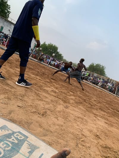 The image shows two men wrestling on a sandy field in Sokoto, Nigeria. One man is wearing blue shorts and a white shirt. The other man is wearing blue shorts. The man in blue shorts is facing the camera, while the man in blue shorts and a white shirt has his back turned to the camera. There are many spectators watching the wrestling match. There is a small building in the background. A person wearing a blue shirt and yellow armband is standing in the foreground, watching the match. There are sandbags and a blue and white canvas on the ground. The sky is clear and blue. The ground is sandy.