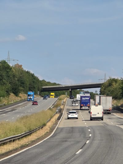A highway overpass spans a multi-lane road in Krunkel, Germany, on a clear day. The road is lined with trees and grass, and power lines are visible in the distance. Several vehicles, including trucks and cars, are traveling on the highway. The traffic is light, and the weather appears to be good.