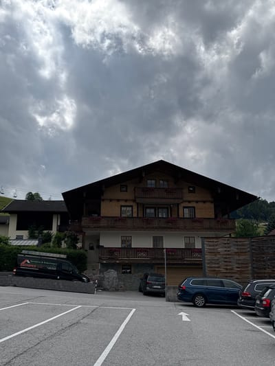 A dark, overcast sky looms over a multi-story chalet-style building made of wood and stucco. The building features multiple balconies with wooden railings adorned with red flowers. A black van with "DACHS PEZI" and "Spenglerei & Bedachungen" written on its side is parked on a sloped area in front of a stone wall. Several cars are parked in a paved lot in the foreground, including a dark blue station wagon and a black hatchback. To the left of the chalet, another building with a dark roof is visible, and further in the distance, ski lift poles ascend a green hillside. The scene appears to be a residential or commercial area in a mountainous region.