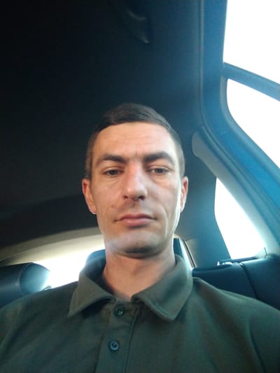 A man is taking a selfie inside a car. The man is in his 30s, with short brown hair and a light complexion. He is wearing a dark green polo shirt. The background of the image shows the interior of the car, including the headrest of a seat and the car's windows. The image appears to have been taken during the day, as evidenced by the bright light coming through the windows. There are no specific landmarks or indicators that would pinpoint the location to Bene, Ukraine.