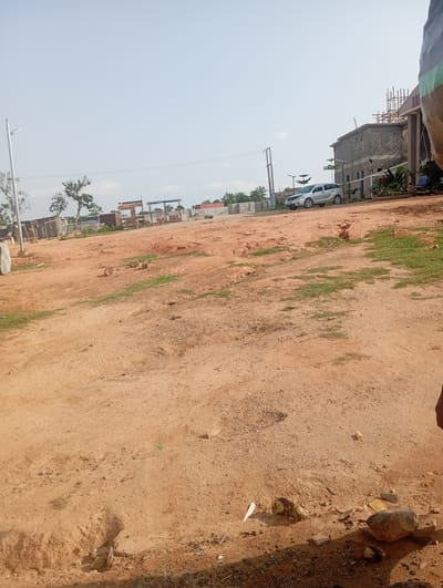 The image depicts an outdoor scene in Tasha, Nigeria. The foreground is dominated by a dusty, reddish-brown dirt ground with some scattered rocks and debris. Patches of dry grass are visible in places. In the middle ground, there are several makeshift structures, possibly market stalls or temporary shelters, constructed from wood and corrugated metal. Further in the background, a modern, grey stone building is under construction, with scaffolding visible on its upper levels. A silver SUV is parked in front of this building. A motorcycle with a rider is also visible near the building. A street lamp stands on the left side of the image. The sky is clear and bright, suggesting it is daytime, likely mid-morning or afternoon. The overall impression is of a developing area, possibly a construction site or a marketplace on the outskirts of a town. There is no visible text in the image.