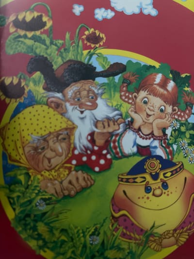 This is an image of a colorful illustration featuring several cartoon characters, likely from a children's story or folk tale. The subjects include a young girl with red braided pigtails, freckles, and a mischievous grin. She is wearing a traditional-style dress with red and green embroidery. Beside her are two older individuals, a man and a woman, who appear to be gnomes or dwarves. The man has a long white beard, is wearing a dark hat and a red polka-dot shirt, and has a large, jovial smile. The woman is wearing a yellow polka-dot headscarf and also has a warm, grandmotherly expression. They are all situated in a grassy, outdoor setting with sunflowers and small white flowers visible. The overall mood is cheerful and whimsical. To the right of the scene is a large, decorative object, possibly a pot or a shield, adorned with blue and yellow patterns. The image appears to be a painting or drawing with a slightly rustic, storybook aesthetic. There is no visible text in the image. Given the provided location context of a rural settlement in Russia, the imagery might be inspired by Slavic folklore or fairy tales. The time of day and weather are not explicitly indicated but the bright lighting suggests daytime and fair weather.