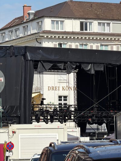 The image shows a large outdoor stage setup in front of a white building with a dark tiled roof. The stage is covered with black fabric and appears to be equipped with lighting rigs and speakers. A partial view of the building behind the stage reveals windows and architectural details. Text on the building reads "DREI KÖNIG." In the foreground, a white container unit is visible, with a red electrical socket and some wiring. A traffic sign with a blue circle, red cross, and white arrow pointing left is mounted on a pole to the left of the container. The ground is paved, and parts of vehicles are visible in the lower right corner. The overall scene suggests an event or performance setup in an urban environment. The text "DREI KÖNIG" likely refers to a hotel or business named "Drei Könige," and the context of a stage setup suggests a public event. The presence of a traffic sign indicates a street or public space.
