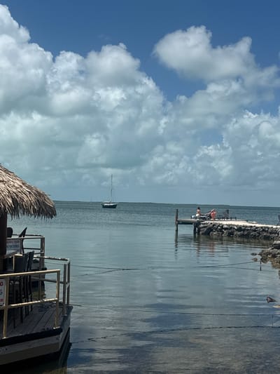 The image depicts a coastal scene in Key Largo, Florida, United States. The foreground features a wooden deck with a thatched-roof structure partially visible, along with patio furniture. To the right, a pier extends into the calm water. Two people are sitting on the pier. A sailboat is anchored in the distance. The sky is filled with scattered clouds.