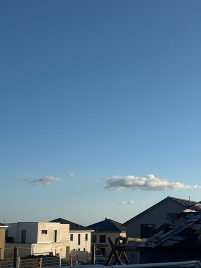 The image depicts a residential area in Pirmasens, Germany, under a clear, light blue sky with scattered white cumulus clouds. The scene is illuminated by natural light, suggesting late afternoon or early evening.
The upper two-thirds of the frame are dominated by the expanse of the blue sky, featuring two small, dark, indistinct specks, possibly birds or insects, in the upper portion. Towards the horizon, several small to medium-sized cumulus clouds are visible.
The lower third of the image contains multiple houses and foreground structures. In the immediate foreground on the right, a dark, partially obscured roof structure is visible, featuring weathered wooden elements that appear to be part of an external fixture or a smaller, detached structure. Below this, metal railings or pipes are visible horizontally.
To the mid-right, a modern house with a steep, dark-tiled gabled roof and light-colored walls stands. A single, dark, rectangular window is positioned in the upper wall section.
In the mid-ground, slightly left of center, a distinct modern house with a cubic, minimalist design is present. It features light-toned, possibly stucco or concrete, walls and flat roof sections. A smaller, projecting cubic element on its front facade contains a small, rectangular window and a taller, narrow vertical opening. To its right, another tall, narrow window is visible. An external air conditioning unit is mounted on the left side of this house. Behind it, further structures with dark, sloped roofs are partially visible. On the far left horizon, a faint outline of a wind turbine can be discerned.
No people are visible in the scene. The overall environment suggests a contemporary suburban or exurban development.